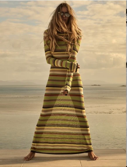 A tall, fair-skinned woman wearing sunglasses and a striped maxi dress poses barefoot on a sunlit stone patio with a sea view.