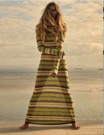 A tall, fair-skinned woman wearing sunglasses and a striped maxi dress poses barefoot on a sunlit stone patio with a sea view.