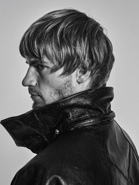 Black and white portrait of an adult male with blond hair and a beard wearing a black leather jacket against a neutral background.