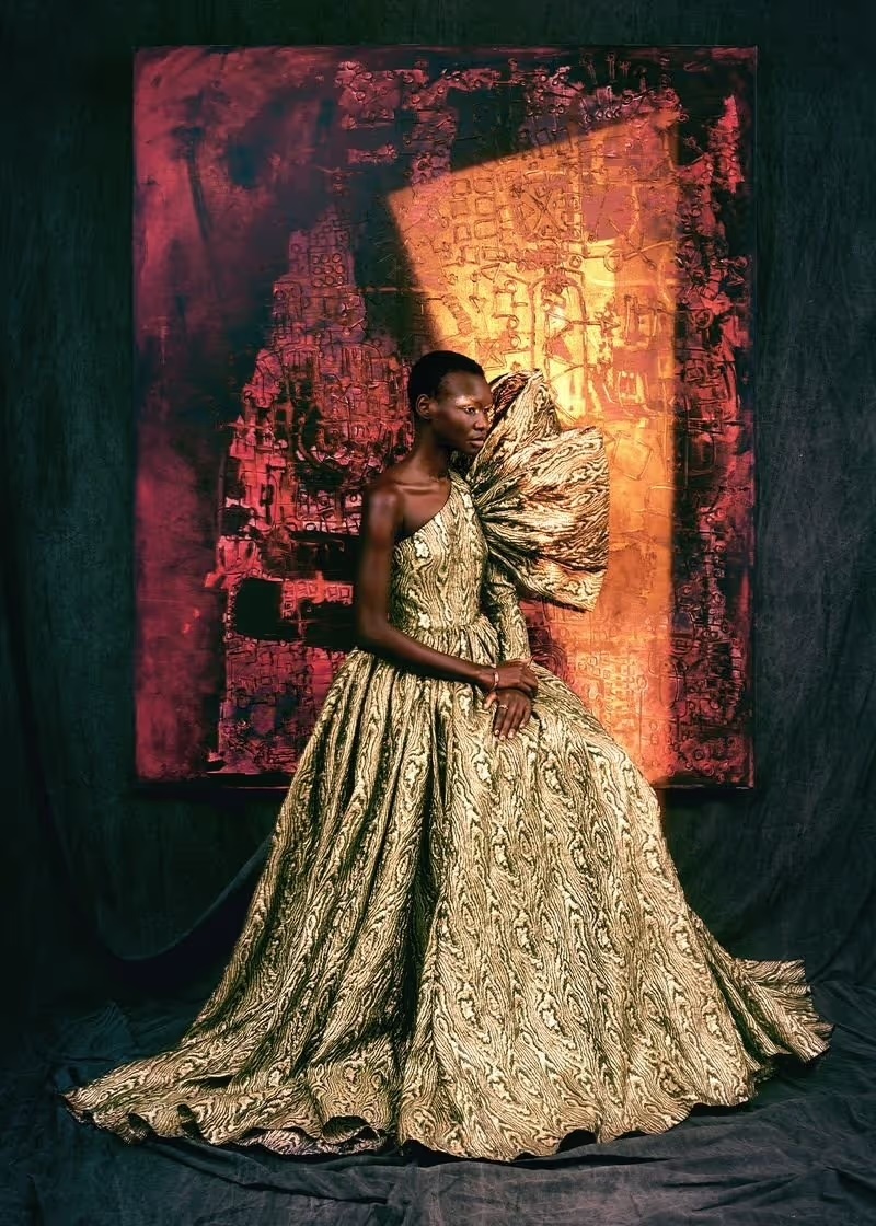 An elegant Black woman poses in a patterned gown with a large shoulder detail, sitting in front of a textured art piece, beauty shot.