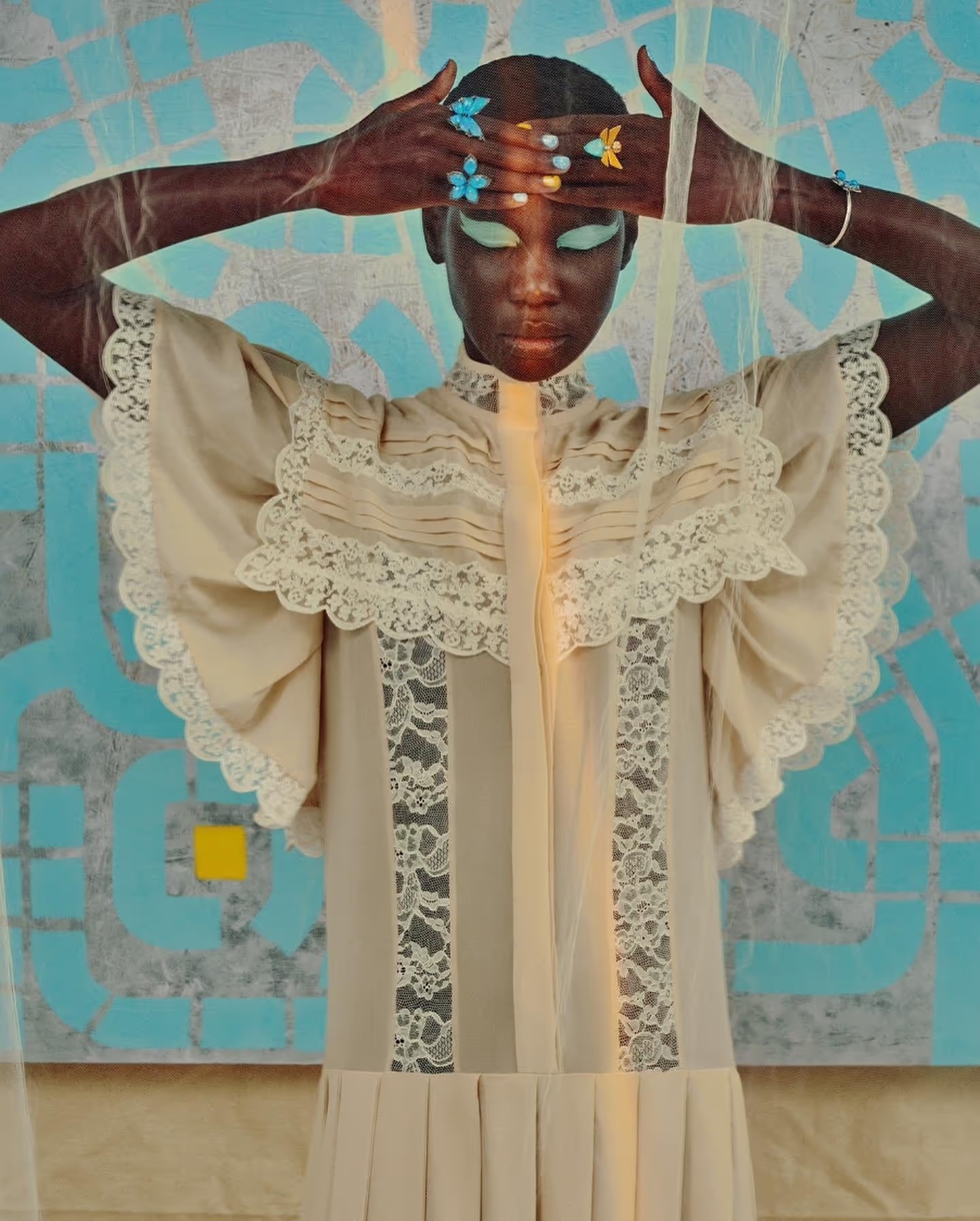 Fashion model wearing a light beige vintage dress with lace detail against a blue patterned backdrop, wearing floral rings, studio lit.