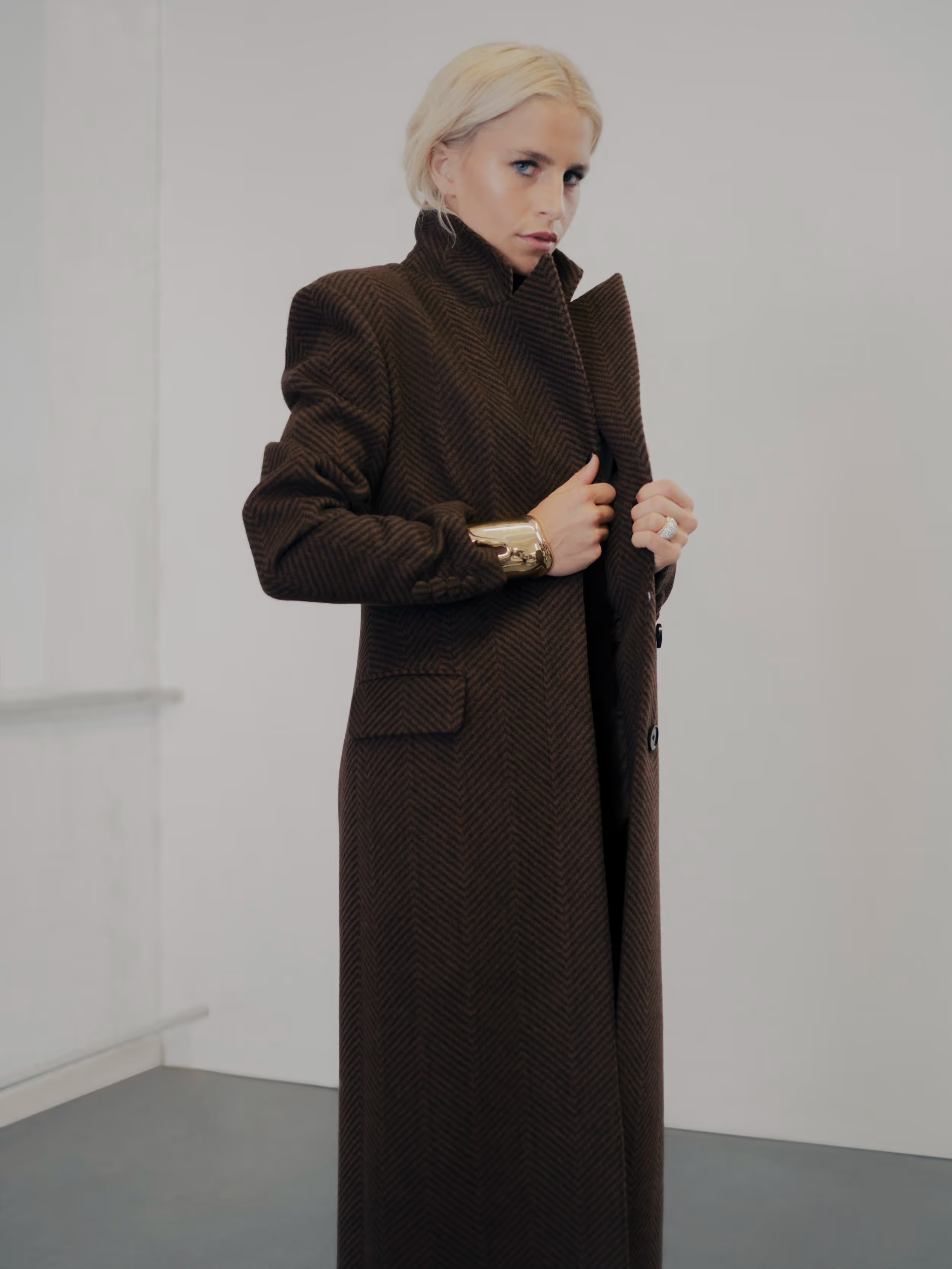 Elegant woman in a long brown coat stands in a minimalist setting, showcasing fashion and style against a plain white backdrop.