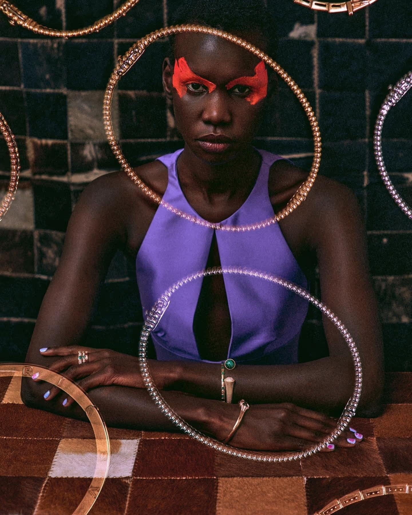 A striking portrait of a woman with face paint wearing a purple top surrounded by circular necklaces against a tiled wall.