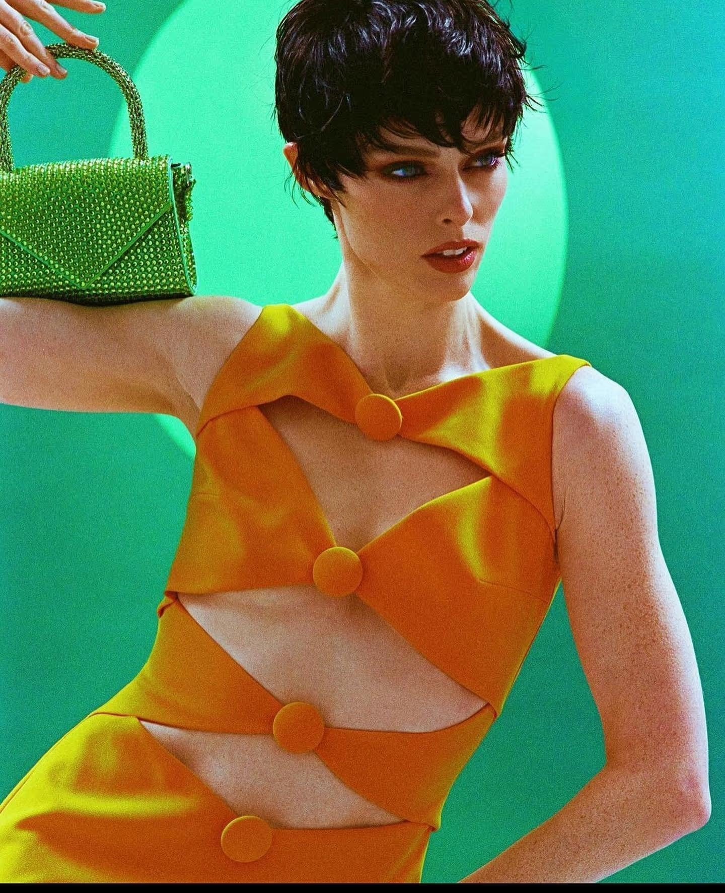 A woman poses with a bright green, studded handbag in a studio shot, wearing an orange dress with geometric cutout details against a green backdrop.