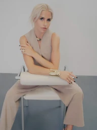 Blonde woman wearing beige sleeveless jumpsuit and gold cuff bracelet seated on a white chair against a grey studio backdrop