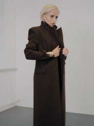 Model wearing a structured long brown herringbone coat with a high collar and a wide gold cuff bracelet against a plain wall.