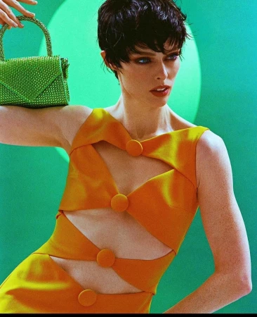 A short-haired woman wearing a yellow cutout dress holding a green embellished handbag against a vibrant green backdrop