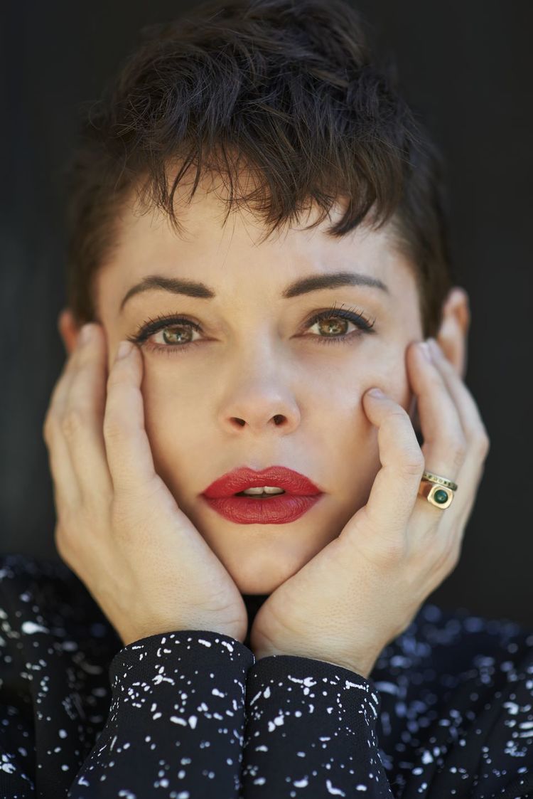 Close-up portrait of a woman with short dark hair and red lipstick, holding her hands to her face against a dark background.