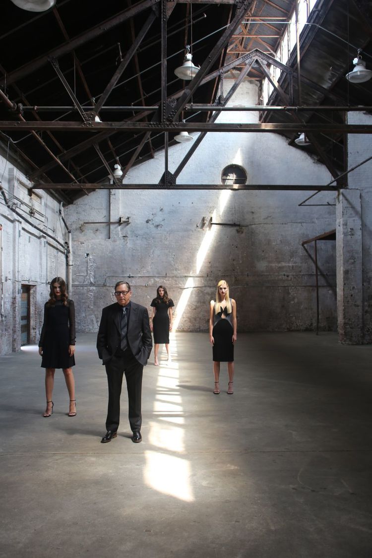 Four adults in formal black clothing pose in an empty warehouse, featuring exposed beams, concrete walls, and bright overhead lighting.
