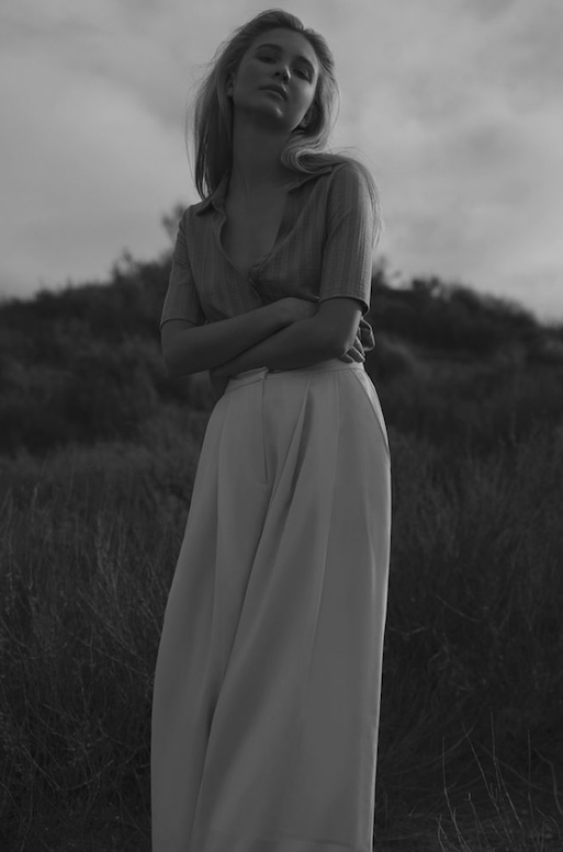 Black and white portrait of a young adult woman with long blonde hair, wearing a collared shirt and wide-leg pants, standing outdoors.
