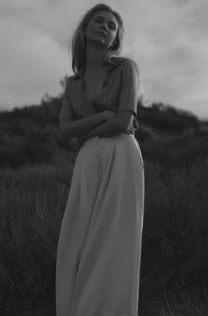 Black and white portrait of a young adult woman with long blonde hair, wearing a collared shirt and wide-leg pants, standing outdoors.