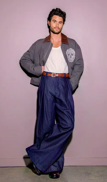 A full-length portrait of an adult male wearing a jacket, tank top, belt, and wide-leg pants. He is posed in a photo studio.