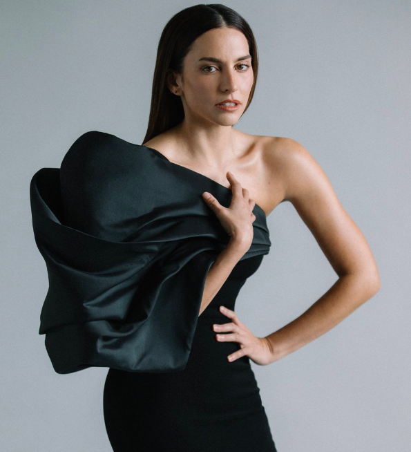 Elegant brunette poses in a black strapless dress with a large satin rosette detail, set against a neutral studio backdrop, portrait orientation.