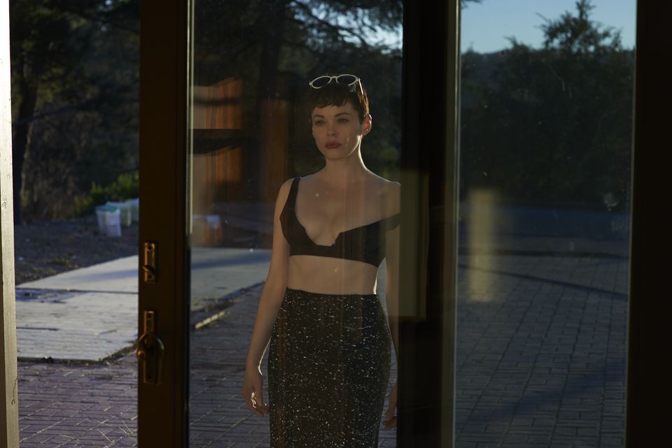 Elegant woman with pixie haircut in black bra top and pencil skirt stands framed by patio doors against trees and bright sunlight.