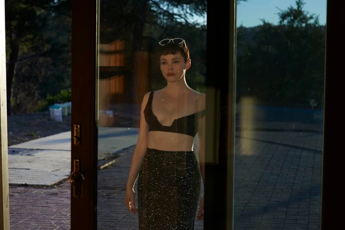 Woman with short dark hair wearing a black crop top and dark skirt standing behind glass doors with trees outside