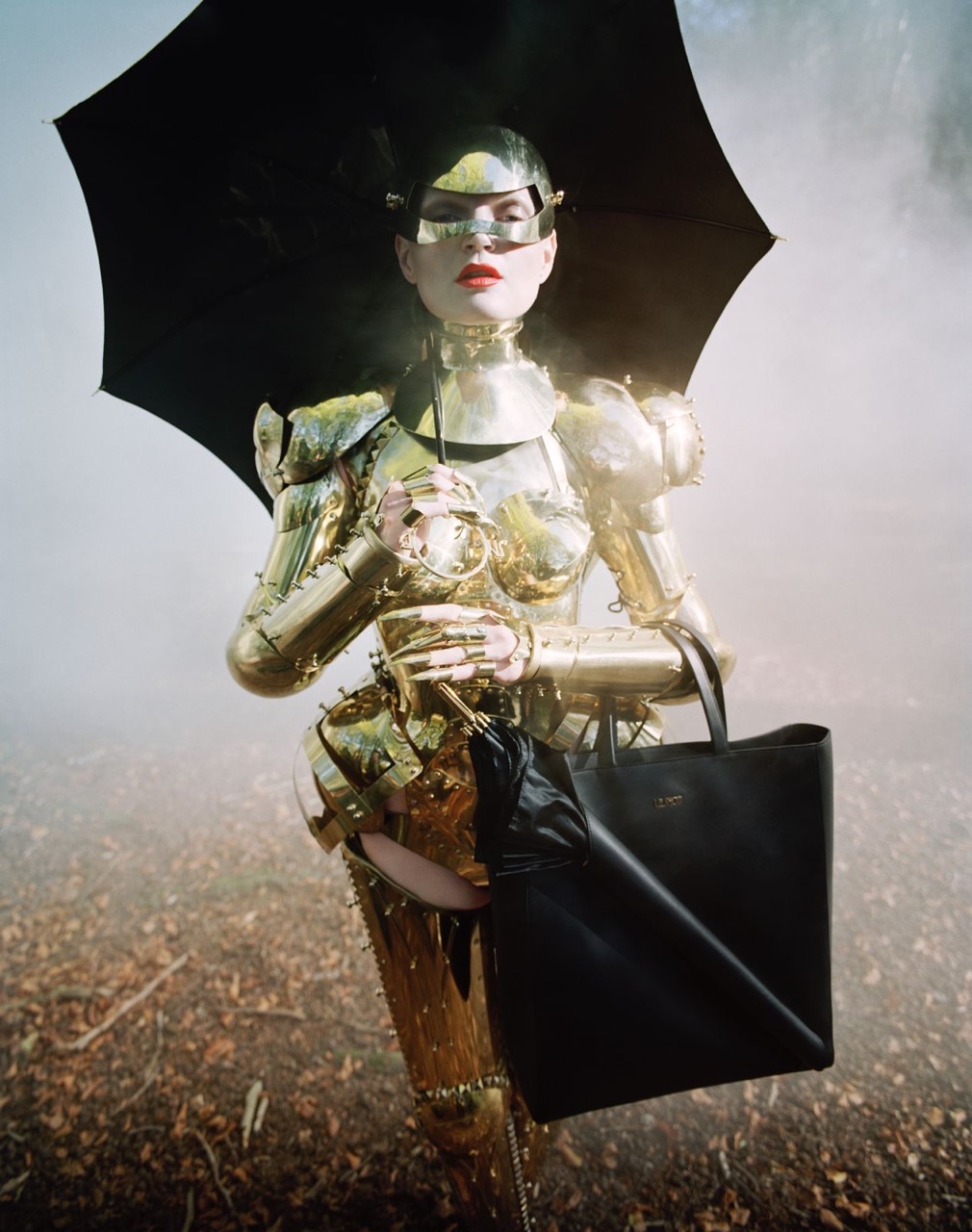 Woman in gold armor, holding umbrella and handbag, poses in a foggy forest. Fashion photography with dramatic lighting and unique style.