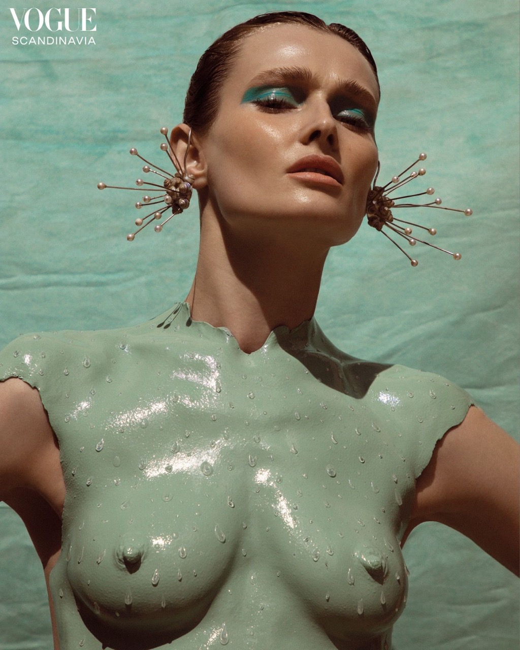 A nude female fashion model is covered in glossy green paste and large jeweled earrings, posing against a mottled green background in studio light.