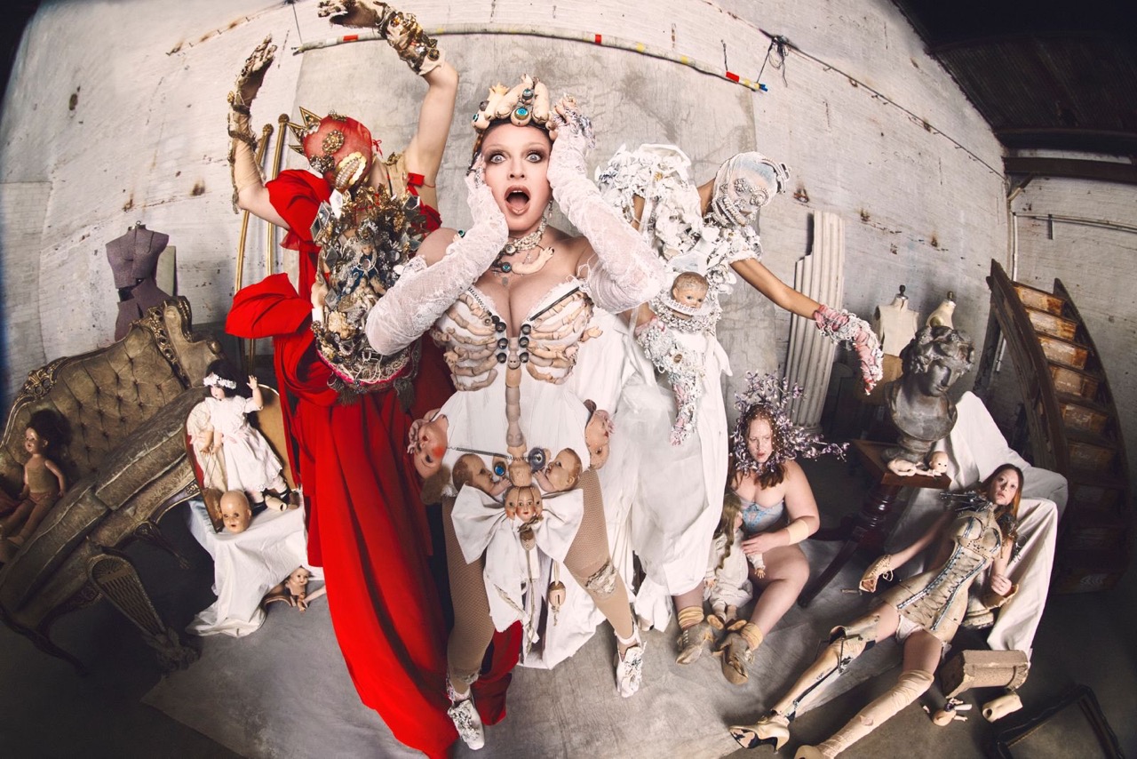 Visually striking portrait features several people in ornate costumes, theatrical makeup, and surreal doll-like props against a textured wall.