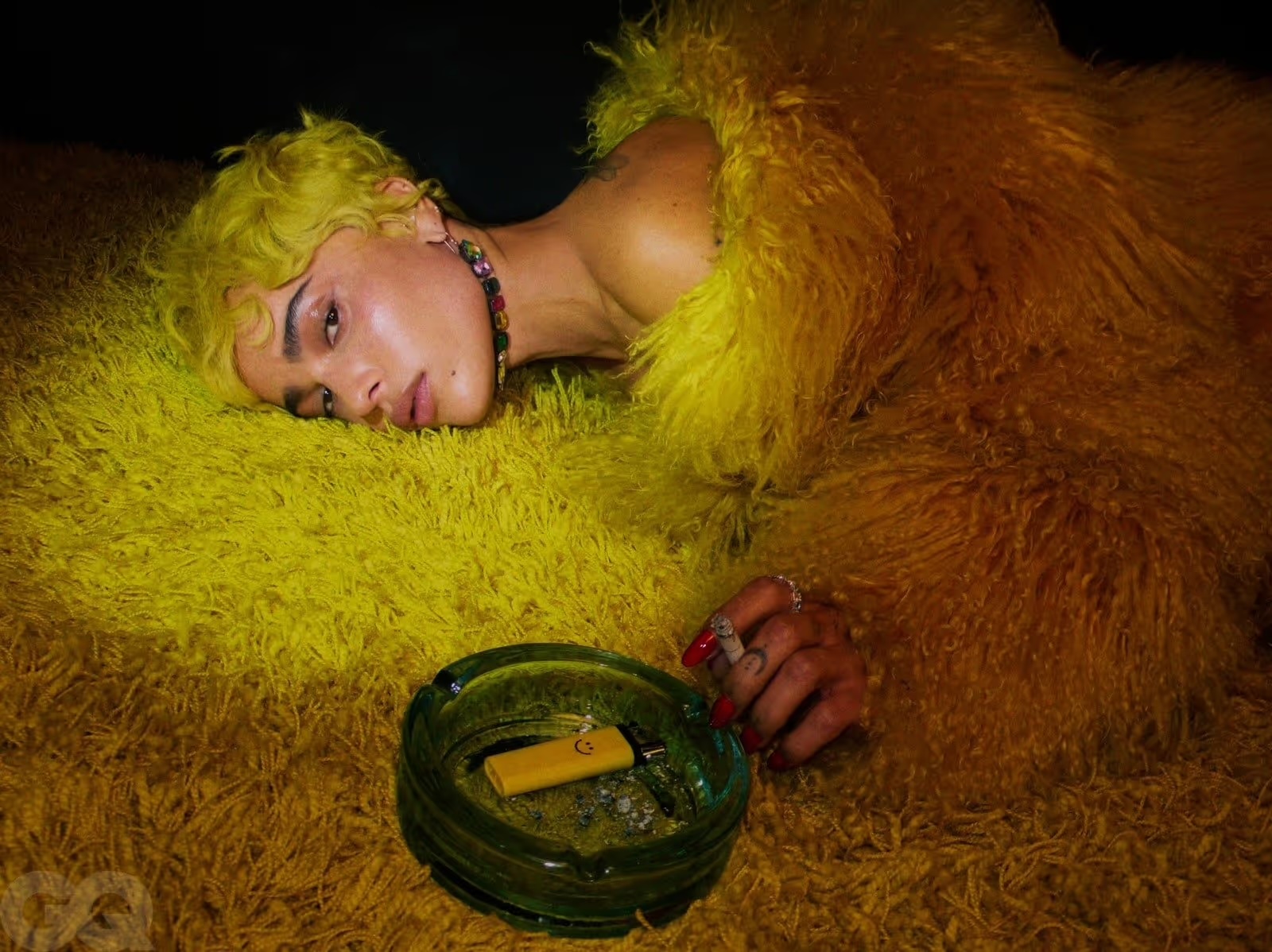 Portrait of a fair-skinned young adult with yellow hair and a colorful necklace, lying on a yellow, shaggy rug, wearing a furry coat.