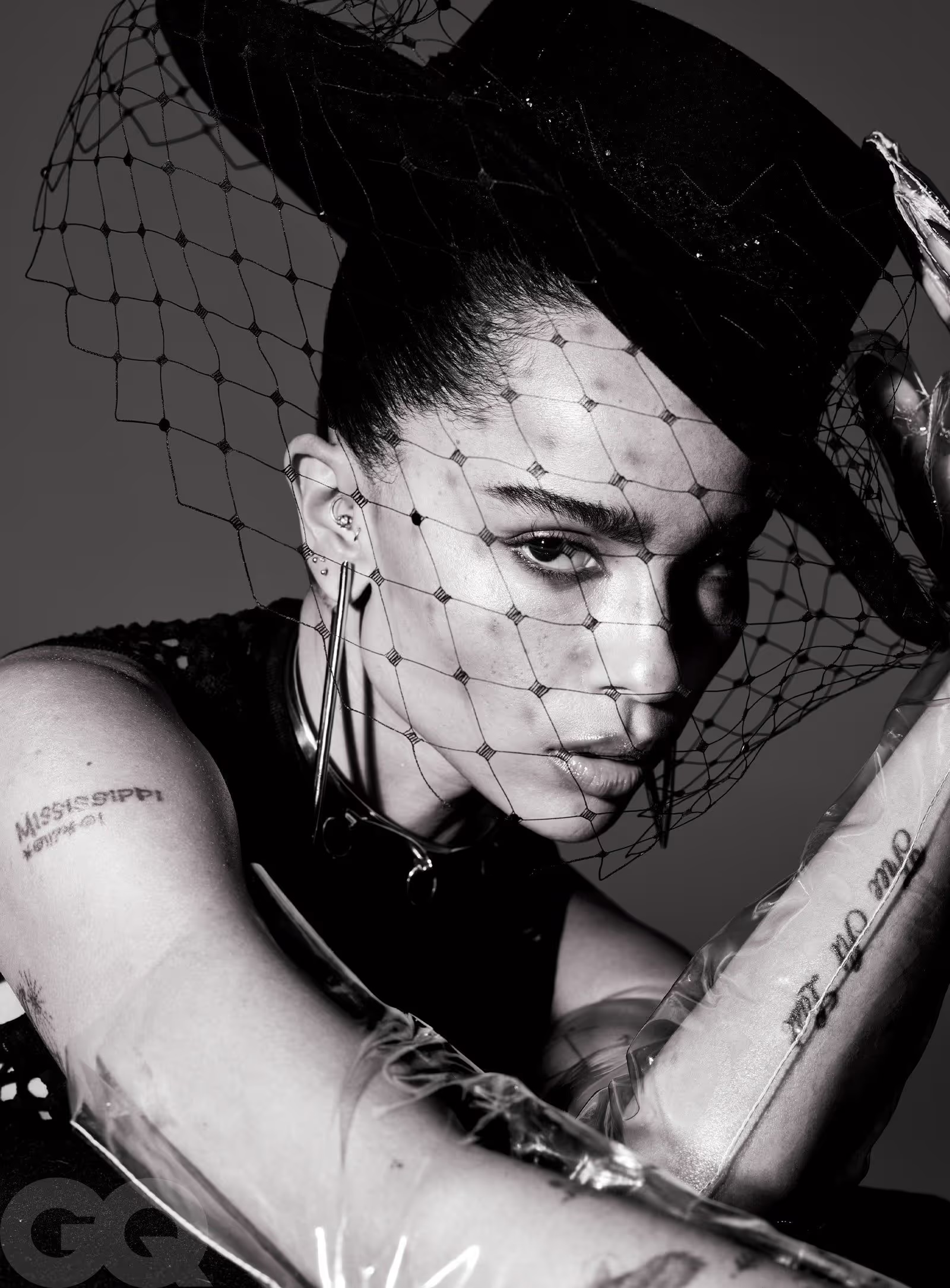 Monochrome studio portrait of a woman wearing a black veiled hat and transparent plastic gloves, posing against a dark grey backdrop.