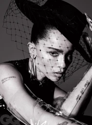 Black and white portrait of a woman wearing a dark hat with a mesh veil, drop earrings, and sheer gloves with visible tattoos.
