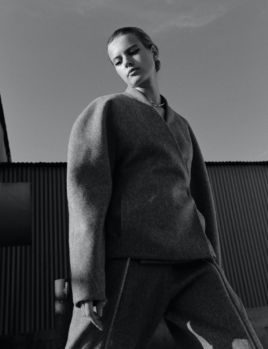 Black and white shot of a woman in a gray wool suit looking down, set against a metal fence and bright sky. Fashion editorial.