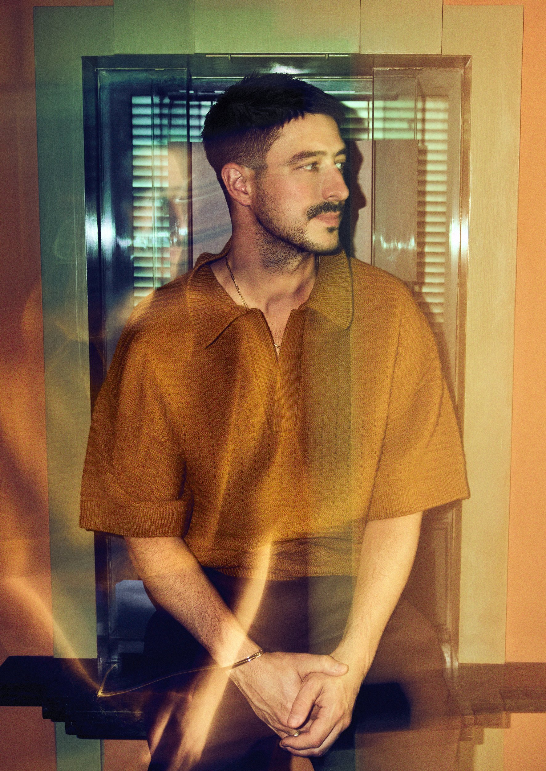 Portrait of a man with a mustache in a short-sleeved polo shirt, standing indoors with soft lighting and geometric background.