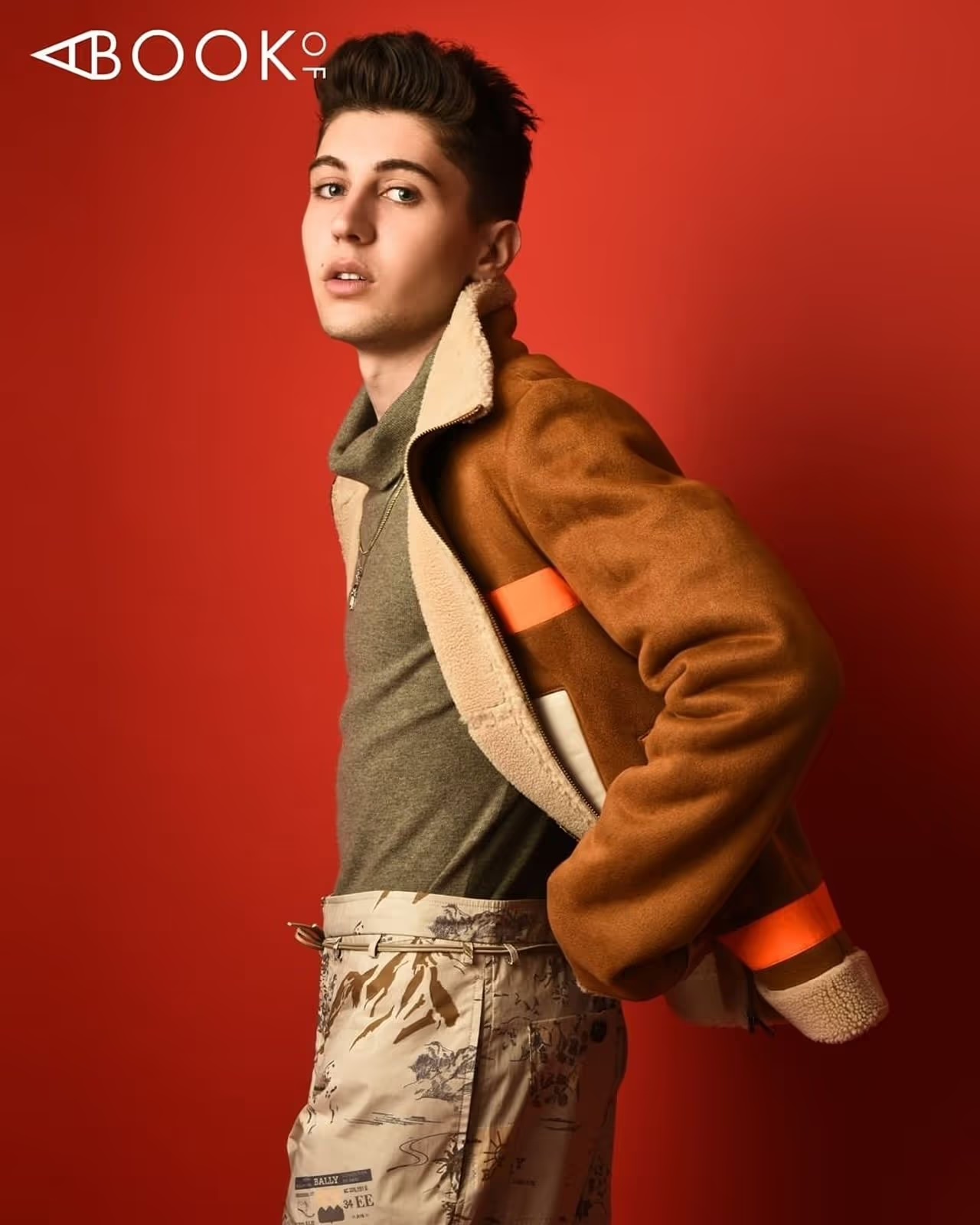 A young adult male model poses indoors against a red backdrop, wearing a brown jacket and printed pants in a fashion editorial shot.