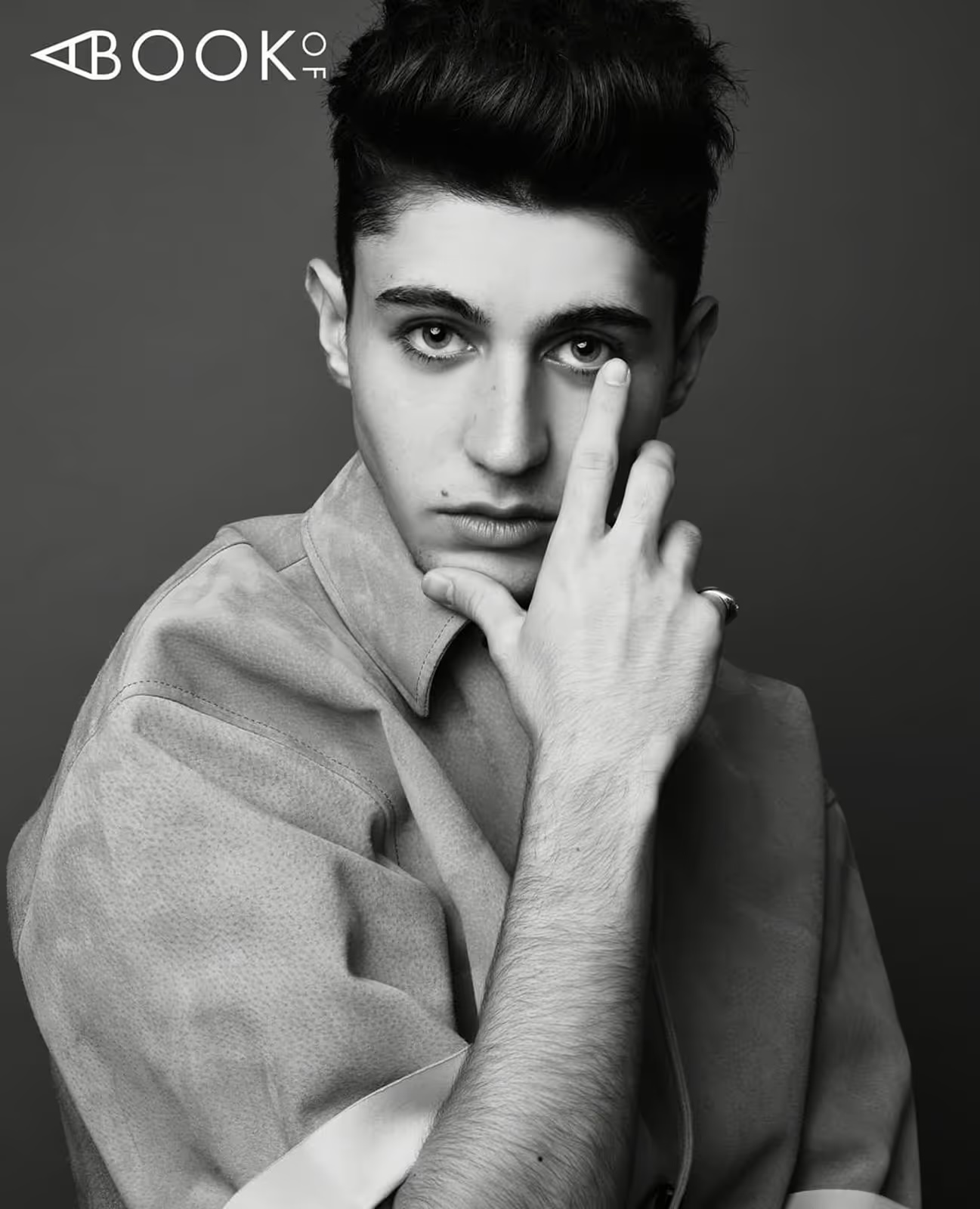 Monochromatic portrait of a young adult male with styled hair, wearing a collared shirt, and hand near his face on a grey background.