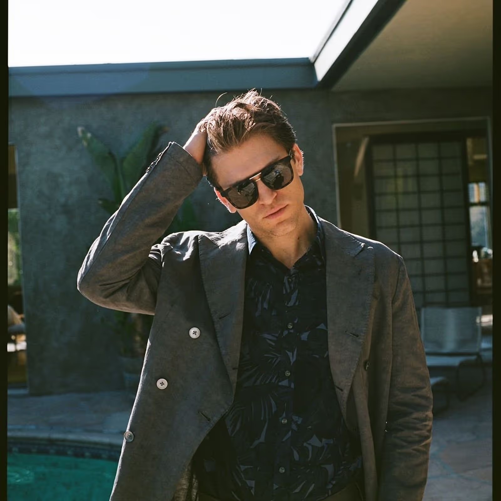 Stylish man in sunglasses poses outdoors in jacket and patterned shirt, hand on head with modern building and pool background.