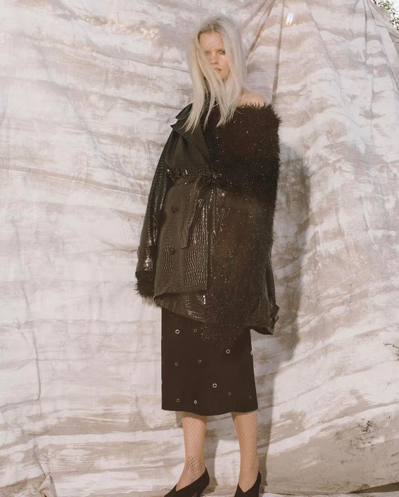 A woman with platinum blonde hair in a shiny black coat and skirt poses against a textured background in a fashion-forward editorial shot.