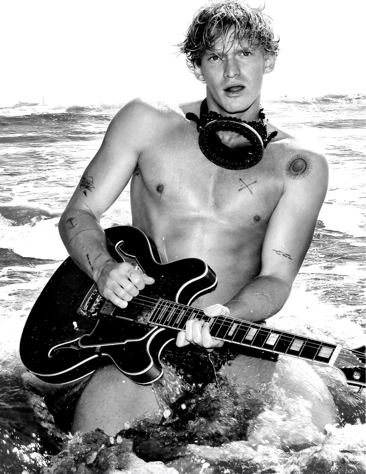 Shirtless man stands in ocean waves playing an electric guitar. He is wearing beaded necklace and has multiple visible tattoos.