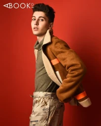 Young man in shearling jacket and patterned trousers posing against a solid red backdrop with Booker magazine logo