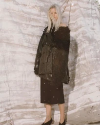 Model with long blonde hair wearing a textured dark coat and skirt standing against a textured backdrop