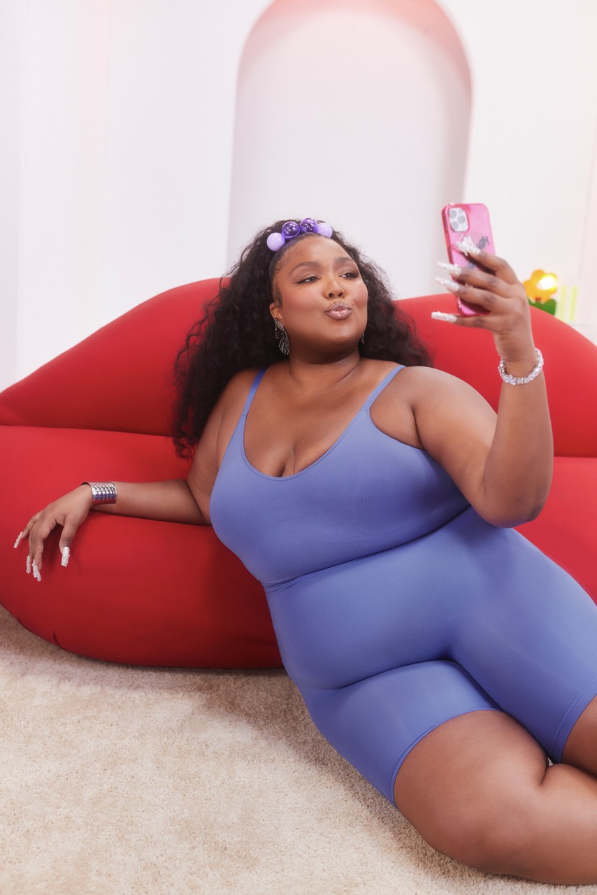 Lizzo poses on a red lips sofa taking a selfie with a pink phone; she is wearing a periwinkle one-piece and purple hair accessories.
