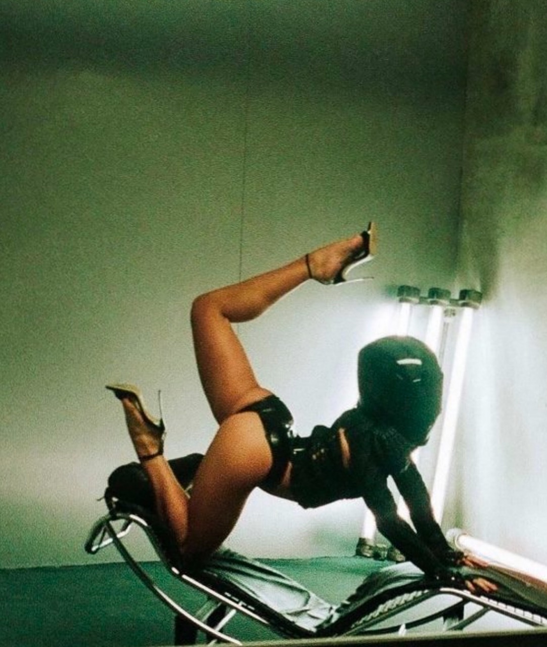 Woman in black latex suit and high heels poses reclining on a chaise lounge chair in a photo studio with fluorescent lighting.
