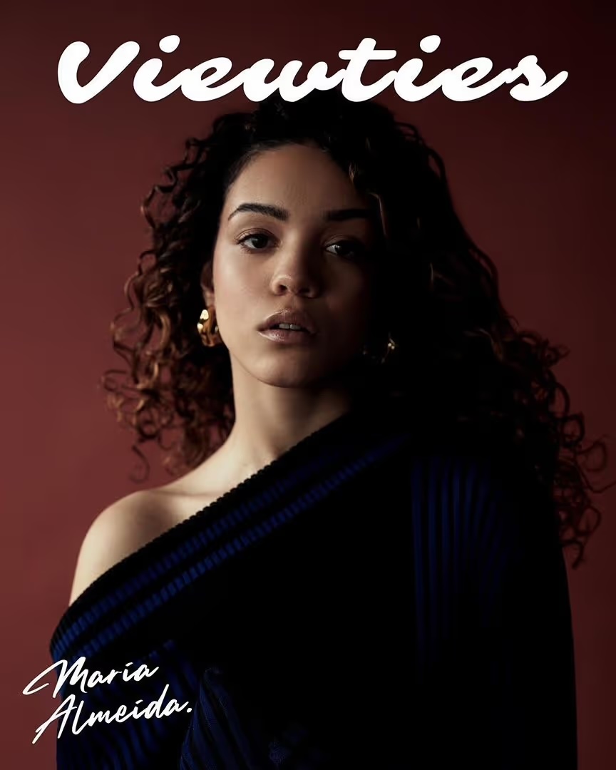 Portrait of a young woman with curly hair, wearing a dark-colored top, posing in front of a red background for a magazine cover.