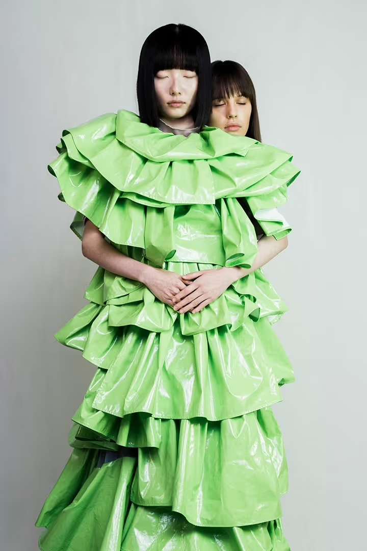 Two women in an avant-garde, shiny lime green tiered gown, eyes closed, standing in front of a light gray background. Fashion editorial.