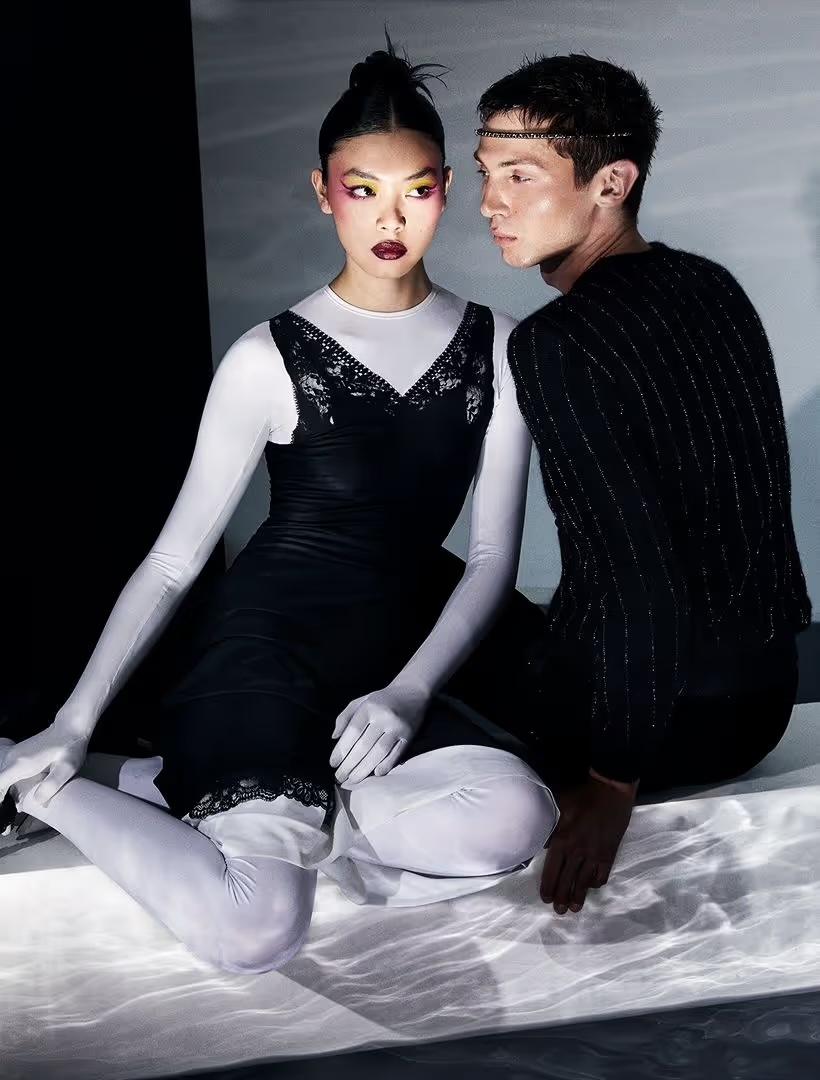 Artistic studio shot of a couple, with the woman in a black dress and the man in a sequined top, kneeling in a stylized indoor setting.