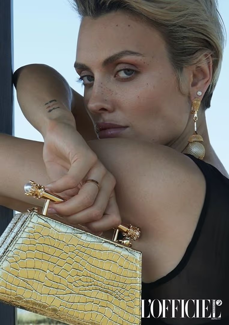 Close-up of blonde woman with golden handbag, accessorized with jewelry and a sheer top, posing with an eye-level angle.