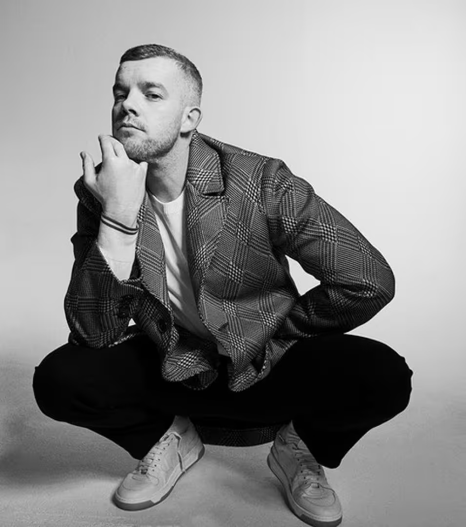 Black and white studio portrait of a man in a plaid blazer and sneakers, crouching with hand to face in a simple photo studio setup.