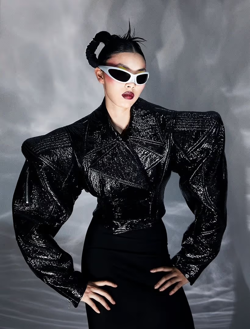 An Asian woman poses indoors wearing sunglasses, a black skirt and a black, high-shouldered jacket against a neutral background.