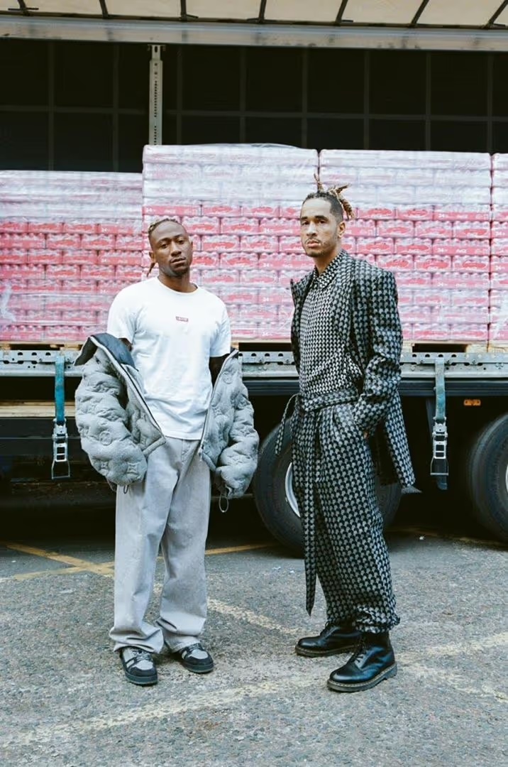 Two adult men stand in fashionable clothing in front of a semi-truck trailer in an urban environment with asphalt ground and diffused light.