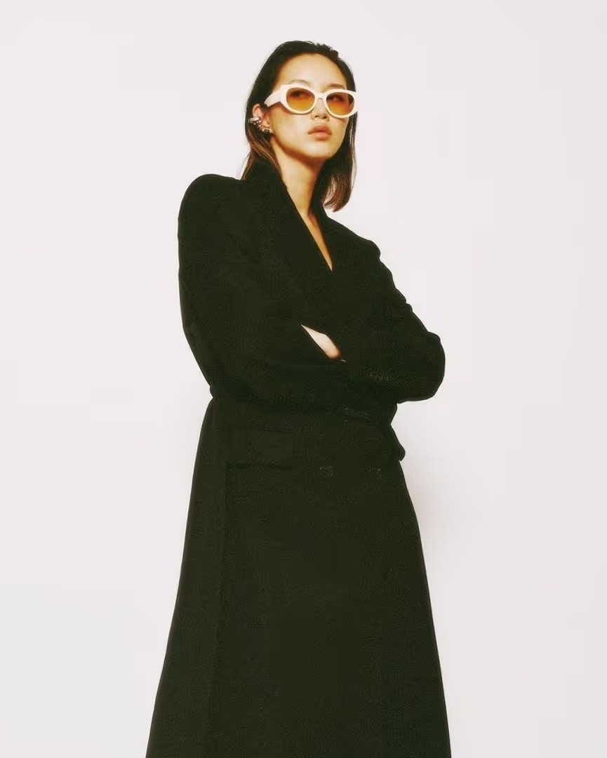Stylish woman in black wrap dress and tinted sunglasses posing in a brightly lit studio. Fashion editorial photography.