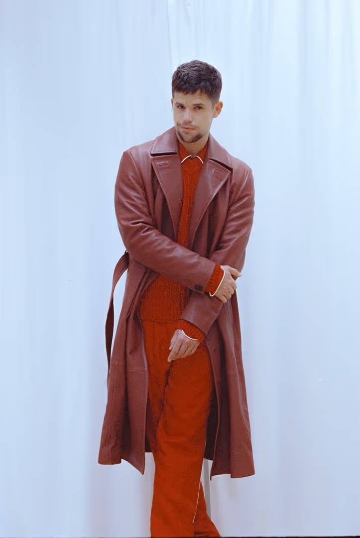 Full-length portrait of a fair-skinned adult male with short dark hair, wearing a red sweater, pants, and long red trench coat indoors.