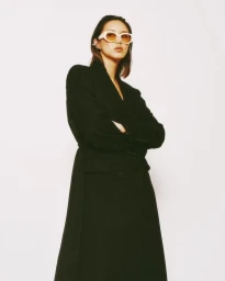 Woman wearing white sunglasses and a long black coat standing against a white seamless background