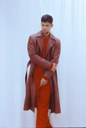 Man wearing a long brown leather coat and red sweater standing against a white seamless background