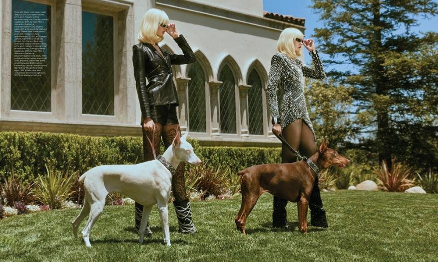 Two fair-skinned adult females with platinum blonde hair pose with a white and a brown Doberman Pinscher on the lawn of a mansion.