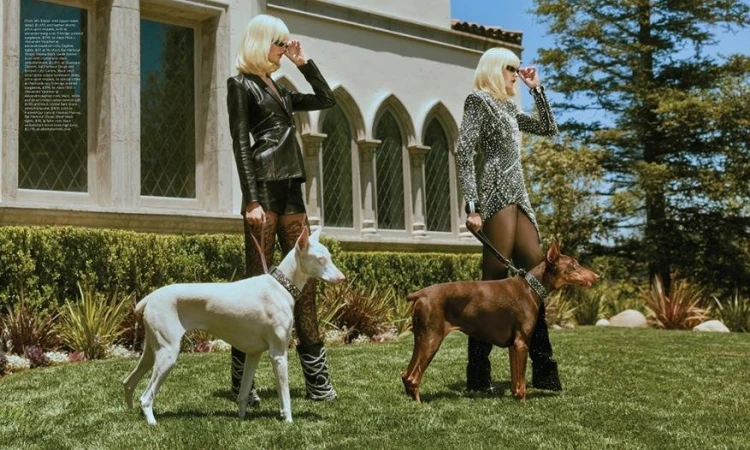 Two fair-skinned adult females with platinum blonde hair pose with a white and a brown Doberman Pinscher on the lawn of a mansion.