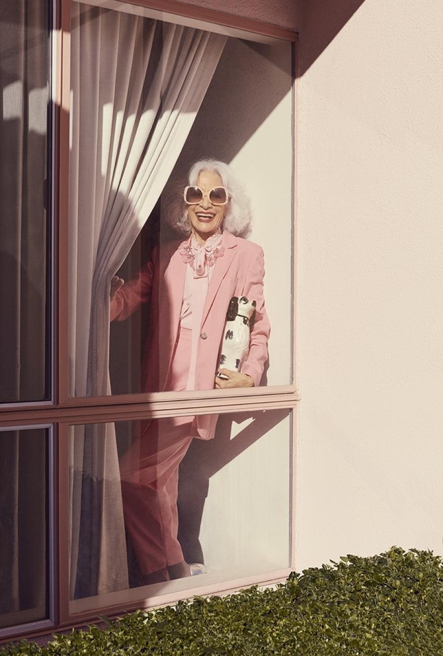 Stylish mature woman in a pink suit and sunglasses holding a patterned vase, framed by a pink wall and window with sheer curtains.